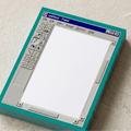Angled view of a thick paper notepad designed to look like a vintage 90s MS Paint window. The pad has a teal border, pixelated tool icons on the left, and a blank white canvas area for writing. The edges of the paper stack show the teal color of the design.